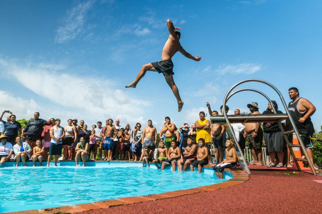 Summer in NZ: A man jumps into a swimming pool as a crowd watches on and cheers. 